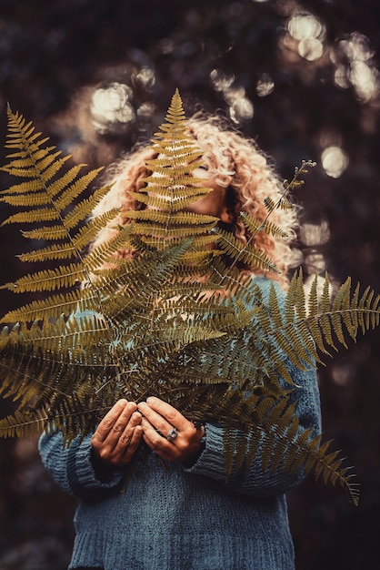 新鮮な壊れやすいシダの葉を持つ青いセーターの幸せな若い女性は、森や公園で植物を残します。植物の葉を保持している満足のいく若い女性。女性は顔の一部を覆って、彼女の手でシダの葉を持っています