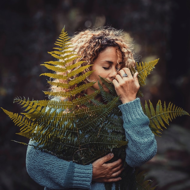 Happy young woman in blue sweater holding fresh fragile fern\
leaves plant in forest or park. satisfied young woman holding plant\
leaves. woman is holding fern leaf in her hands, covering part of\
face