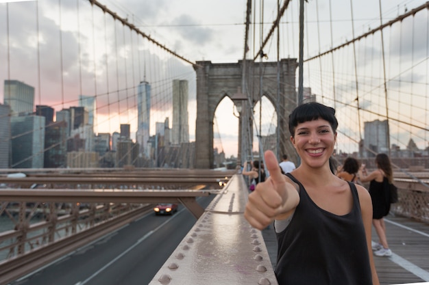 Giovane donna felice sul ponte di brooklyn che mostra i pollici su