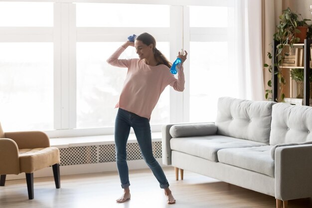 Happy young woman in casual clothes have fun cleaning the home\
holding rag and spray fluid smiling carefree housekeeping female\
lady specialist make house chores dancing with detergent in living\
room