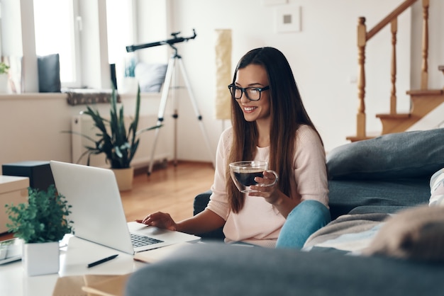 Felice giovane donna in abbigliamento casual che utilizza laptop e sorride mentre riposa a casa at