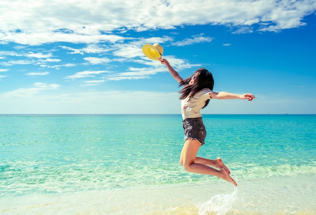 Happy young woman in casual style fashion and straw hat jumping at sand beach.