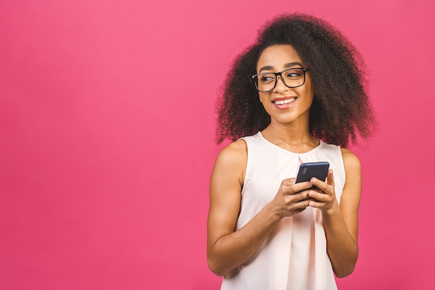 Happy young woman casually dressed standing over pink, holding mobile phone.