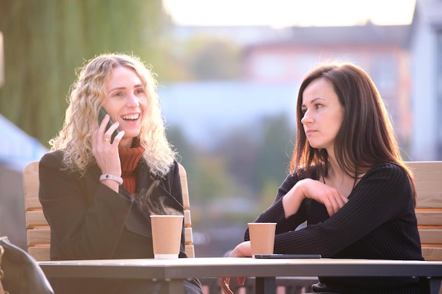 写真 退屈な友人を無視して携帯電話でおしゃべりする幸せな若い女性屋外のストリートカフェに座って、お互いにコミュニケーションに苦労している女性の友人友情問題の概念