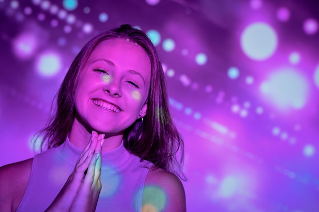 Happy young woman clasping hands and smiling gleefully while making wish during holiday celebration under bright projection of specks of light