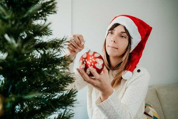 Felice giovane donna che decora il suo albero di natale travestito da un cappello da babbo natale.