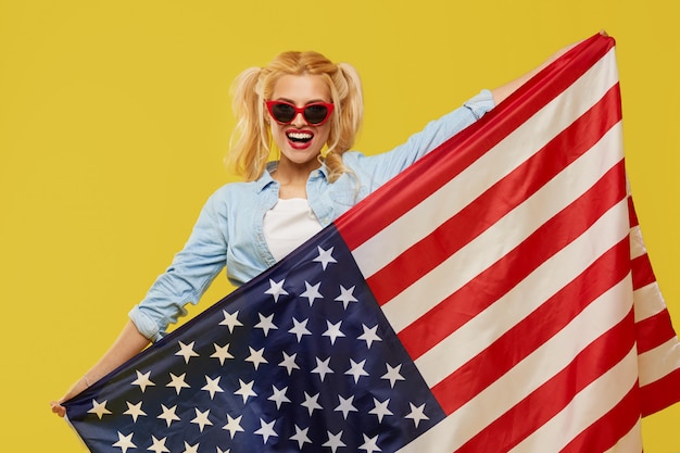Счастливая молодая женщина в джинсовой одежде держит флаг США на желтом фоне