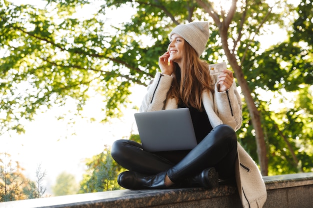 Happy young woman dressed in autumn coat and hat sitting outdoors, using laptop computer, showing plastic credit card