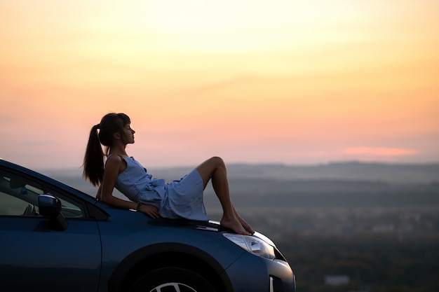 Driver felice della giovane donna in vestito blu che gode della sera di estate calda che pone sul suo cappuccio dell'automobile. concetto di viaggio e vacanza.