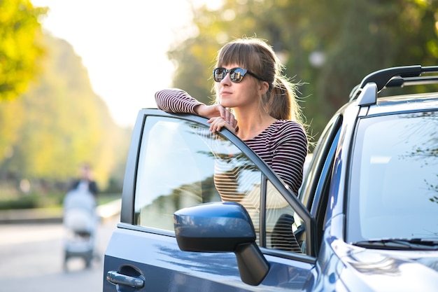 Happy young woman driver enjoying warm summer day standing beside her car on a city street. Travelling and vacation concept.