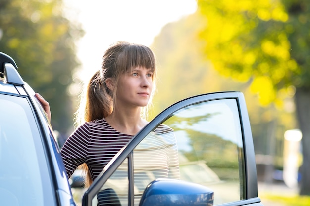 街の通りで彼女の車の横に立って暖かい夏の日を楽しんで幸せな若い女性ドライバー。