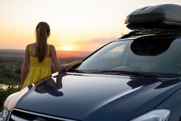 Happy young woman driver in yellow dress enjoying warm summer evening leaning on her car