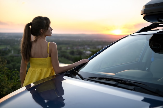 暖かい夏の日を楽しんでいる彼女の車にもたれて黄色のドレスで幸せな若い女性ドライバー。旅行や休暇の概念。