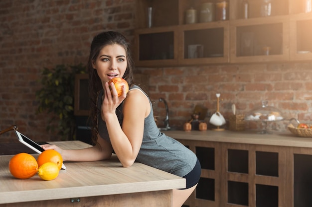 Happy young woman eating fruits and using digital tablet in loft kitchen. Healthy eating, breakfast and dieting concept, copy space