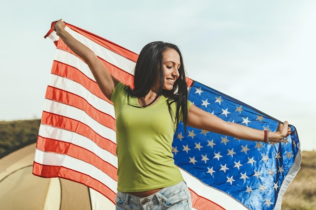 Фото Счастливая молодая женщина наслаждается солнечным днем на природе. она держит американский флаг перед палаткой кемпинга и мечтает о грин-карте.