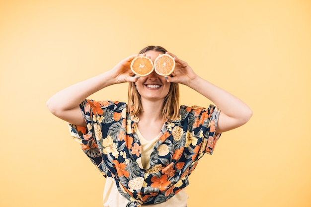 Happy young woman in flowered shirt holding orange halves covering her eyes