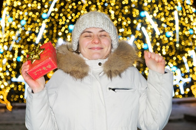 A happy young woman in front of a Christmas tree with bokeh holds a gift and smiles closing her eyes makes a wish