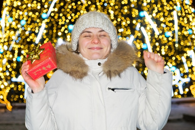 ボケ味のクリスマスツリーの前で幸せな若い女性が贈り物を持って、目を閉じて笑顔が願い事をします