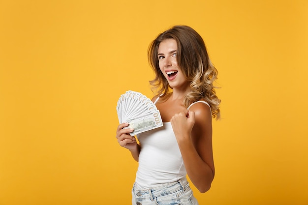 Photo happy young woman girl in light casual clothes posing isolated on yellow background. people lifestyle concept. mock up copy space. holding fan of cash money in dollar banknotes, doing winner gesture.