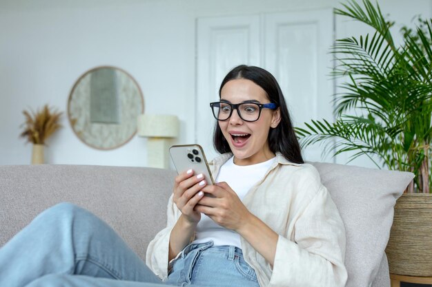 Happy young woman in glasses holding mobile phone celebrating unexpected victory in game won sitting