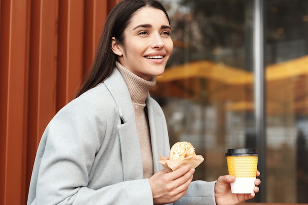 Happy young woman in grey coat eating a donut in a coffee shop.