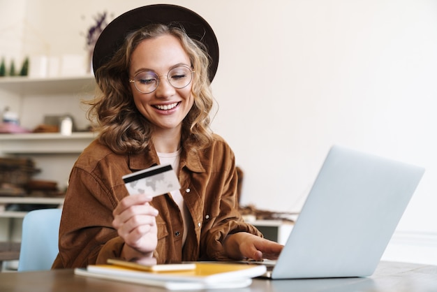 happy young woman in hat working with laptop and holding credit card while sitting at table