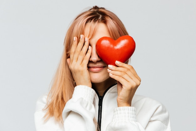 La giovane donna felice tiene il cuore rosso, coprente il suo occhio destro di sua palma, primo piano