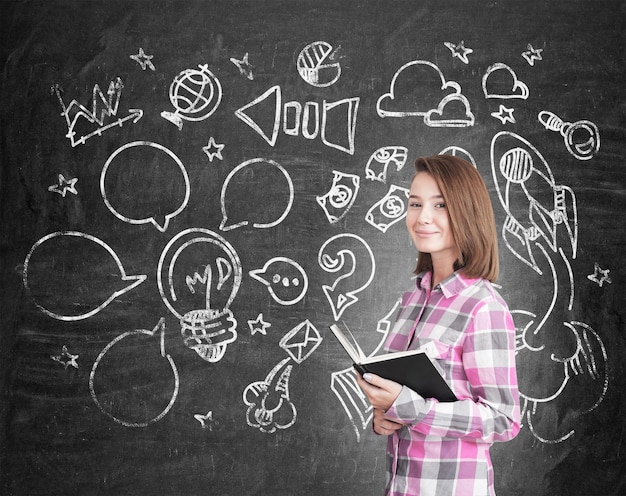 Happy young woman holding a black book and standing near a chalkboard with speech bubbles and arrows. Social media concept