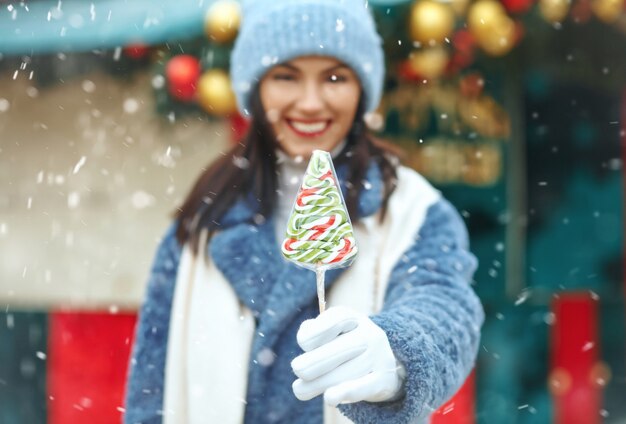 降雪時にクリスマスマーケットでキャンディーを保持している幸せな若い女性