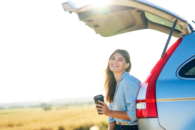 Happy young woman holding coffee cup and sitting on the open trunk of her car in a wheat field at sunset