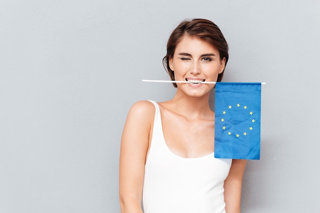 Happy young woman holding european flag in teeth and winking over gray background