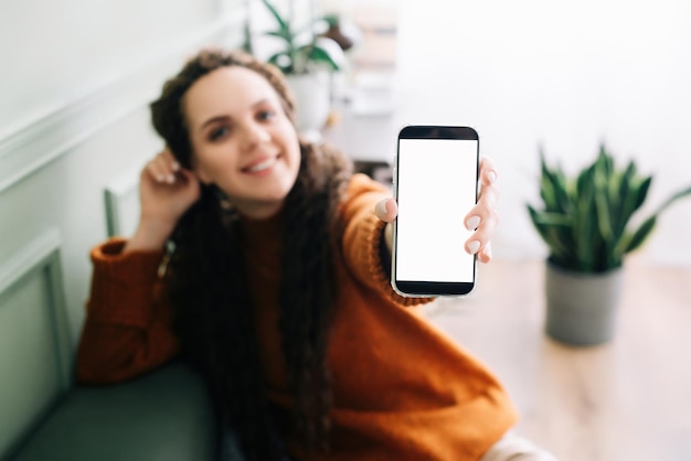 Happy young woman holding mobile phone in hand showing cellphone display presenting big white blank cellular display template mock up smartphone mockup screen for digital application promo ads