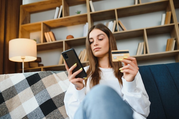 Happy young woman holding phone and credit card sitting on the couch Online shopping