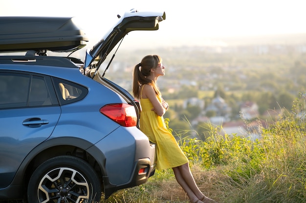 写真 夏の自然の夕日の景色を見ている彼女の車の近くに立っている黄色のドレスを着た幸せな若い女性。旅行と休暇のコンセプト。