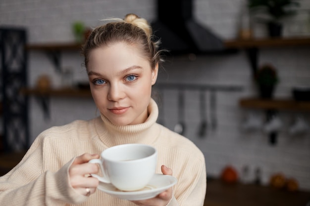 Happy young woman is in a cozy home atmosphere sits at the kitchen table and holds a cup of coffee