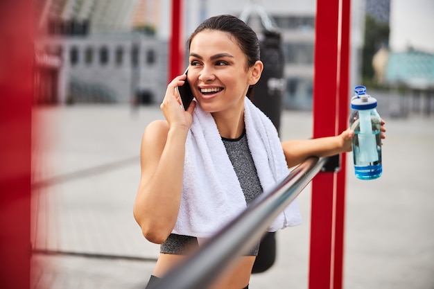 幸せな若い女性は、屋外のスポーツグラウンドでトレーニングをしていて、休憩中に携帯電話で話している