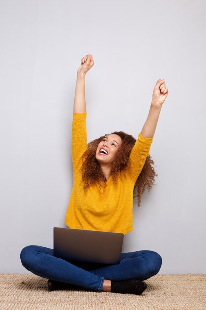 Happy young woman laughing with laptop and arms raised against gray background