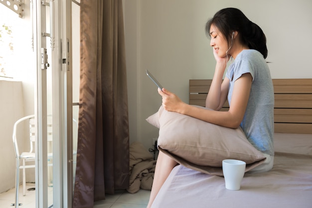 Happy young woman listening music from smart phone with a cup of coffee on the bed.