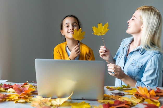 Felice giovane donna e bambina che lavorano online, guardano webinar, podcast sul laptop, conversano a distanza a casa intorno alle foglie d'autunno