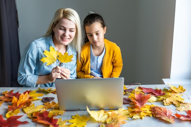 Happy young woman and little girl working online, watching webinar, podcast on laptop, having remote conversation at home around autumn leaves