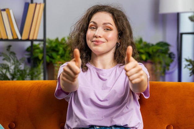 Счастливая молодая женщина с одобрением смотрит в камеру, поднимая большой палец, как положительный знак хороших новостей.