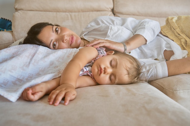 Happy young woman looking at camera while lying near little daughter sleeping peacefully on sofa in living room at daytime