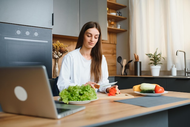 Happy young woman making a salad at the kitchen chopping vegetables looking at laptop computer