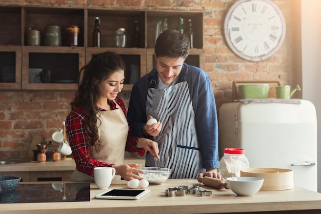 Happy young woman and man baking pie and having fun in loft kitchen. Young family cooking at home, using digital tablet. Mockup for recipe