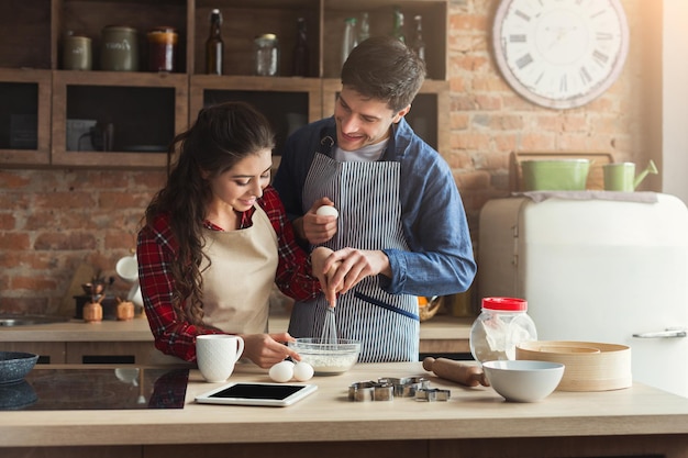 Happy young woman and man baking pie and having fun in loft kitchen. Young family cooking at home, using digital tablet. Mockup for recipe