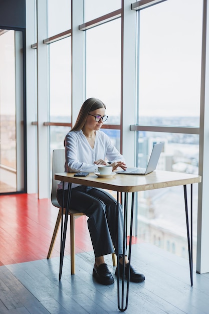 Happy young woman manager in casual attire and glasses smiles and looks into a laptop looking through different information she drinks coffee in a cozy cafe with large windows and a view of the city