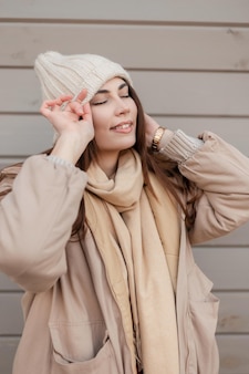 Modello di giovane donna felice con il sorriso in abito invernale di moda con cappello e giacca lavorati a maglia, goditi i momenti all'aperto vicino alla parete di legno