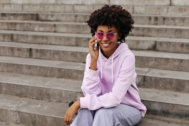 La giovane donna felice in occhiali da sole rosa parla al telefono fuori