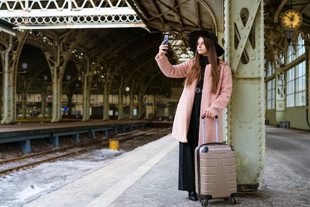 Happy young woman on platform of railway station in pink coat and black hat stands waiting for train...