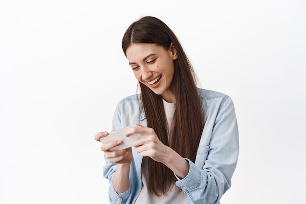 笑って、白い背景に立ってゲームを楽しんで電話画面を見てスマートフォンでビデオゲームをプレイする幸せな若い女性
